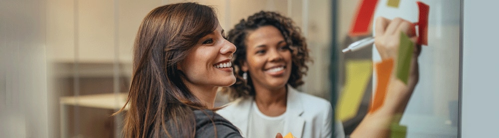 Two consultants write on sticky notes displayed on an office wall