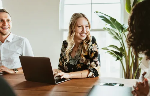 Tre kollegor har ett möte i ett ljust kontor, leende och engagerade i samtal runt ett träbord.