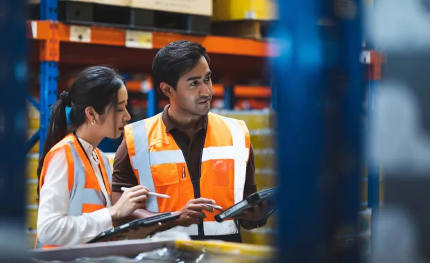 A warehouse worker and manager check stock and inventory using a digital tablet in a retail warehouse full of shelves containing goods